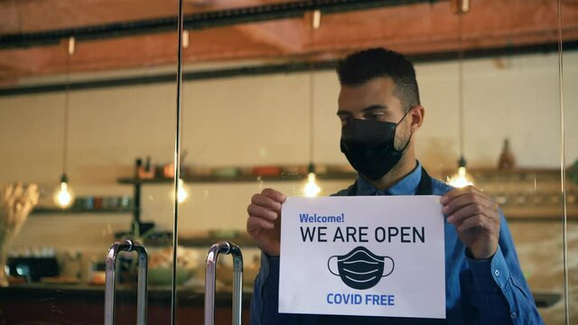 African american waiter with face mask, hangs a Open sign on the front door, after Coronavirus outbreak. Restaurant owner reopens the business. Small business, crisis coronavirus, Keep the distance.