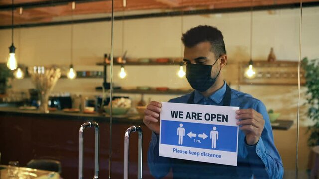 African american waiter with face mask, hangs a Open sign on the front door, after Coronavirus outbreak. Restaurant owner reopens the business. Small business, crisis coronavirus, Keep the distance.