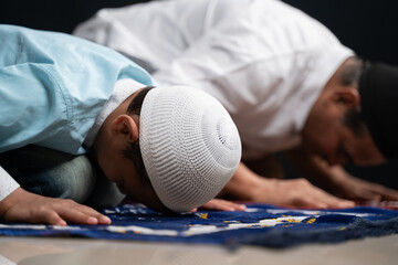 Muslim father and son praying or performing Salah while sitting on Prayer rug and touching head to...