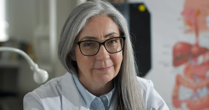 Portrait of smiling woman turning head and looking to camera while sitting in medical office. Close up view of female doctor in glasses and white professional rob. Concept of headshot