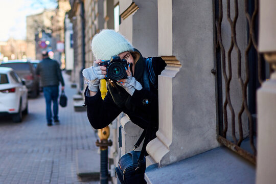 A woman takes pictures hiding behind a wall of a building.