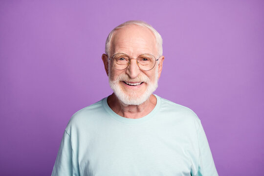 Portrait of smiling optimistic beard pensioner man wear light blue t-shirt eyeglasses isolated on purple color background