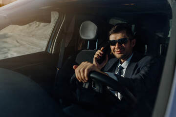 Fototapeta na wymiar businessman in a jacket, shirt, tie and sunglasses is driving a car and talking on a smartphone.