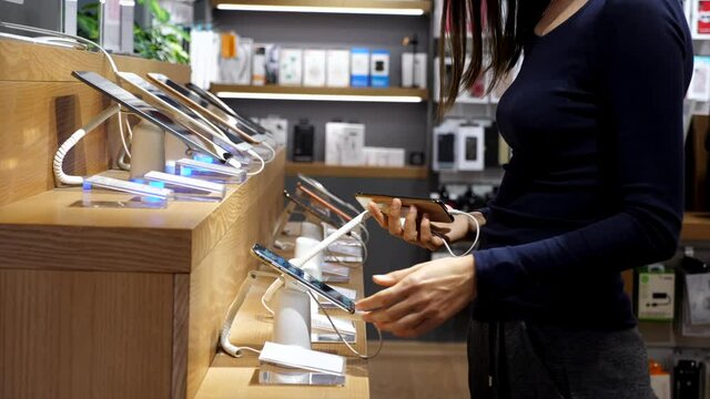 Young woman chooses a new smartphone in an electronics store. Female client is coming to counter, taking two smart phones and comparing it.