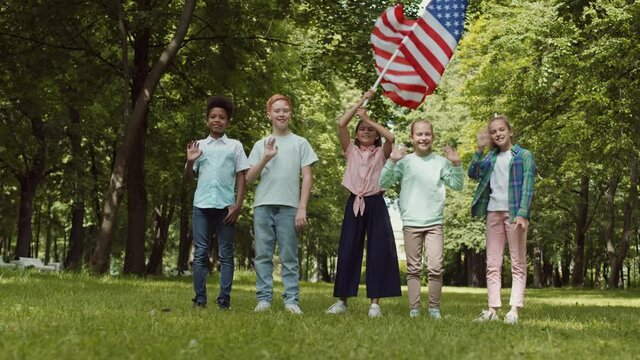 Wide POV shot of four multi-ethnic school male and female children waving, smiling looking on camera, one girl in middle holding fluttering USA flag