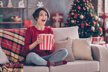 Obraz premium Photo of pretty young lady hold popcorn paper box sitting couch excited open mouth wear red pullover jeans socks indoors