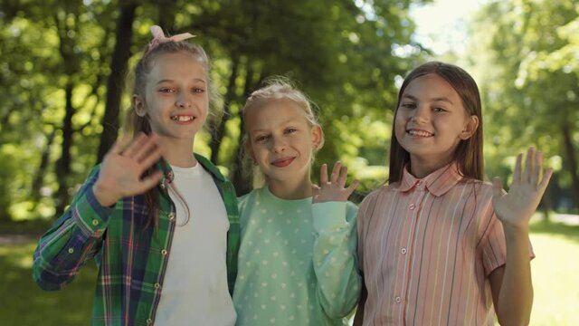 Medium close-up of three Caucasian school girlfriends standing close in park at daytime, looking, smiling and waving on camera