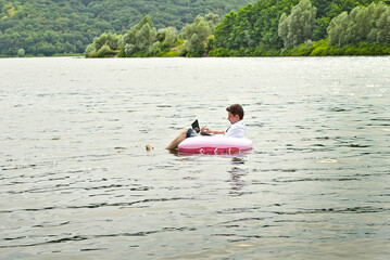Fototapeta na wymiar A man in a shirt on a pink inflatable circle. Guy with a laptop swims in the river. The concept of work during vacation, outsourcing and freelance. Place for text and copy space near businessman.