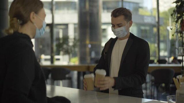 Positive young businessman in Covid-19 face mask receiving takeaway coffee at cafe counter and leaving. Portrait of satisfied customer buying refreshing drink in the morning on coronavirus pandemic.