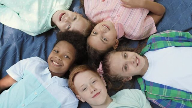 Top Close-up Of Five Multi-ethnic Male And Female School Kids Laying Down On Blue Blanket Head To Head In Circle, Looking And Smiling On Camera