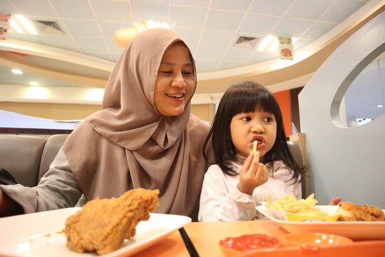 Asian muslim mother and baby girl daughter eating at fast food restaurant, family enjoys fried chicken, burger and potato chips