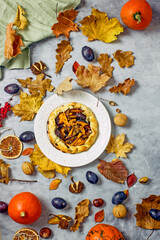 Autumn pie with plums and pumpkin on a light plate surrounded by fallen leaves, acorns, plums, linen napkins. Autumn still life with homemade cakes. Thanksgiving pie Top view, flat lay