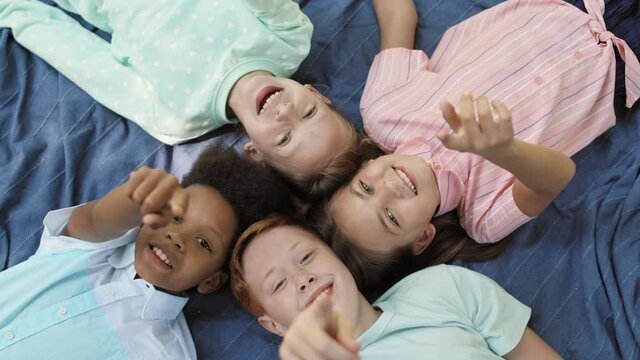 Top Close-up Of Four Caucasian And African School Boys And Girls Laying Down On Blue Blanket Head To Head, Laughing Looking And Pointing Fingers On Camera