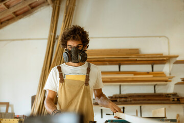 A young man working as carpenter in his wood workshop. Wood worker designing and handcrafting new house furniture using a piece of timber and wearing a full face mask for the dust
