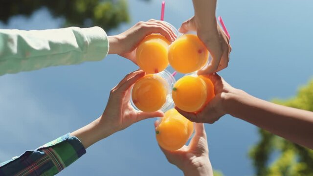 Low angle of blurred blue sky and tree crowns, five unrecognizable hands getting together from out of frame to clink plastic glasses with straws and orange juice