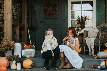 children a boy and a girl with their poodle dog in protective masks on the porch of the backyard decorated with pumpkins in autumn, the concept of the covid-19 pandemic quarantine