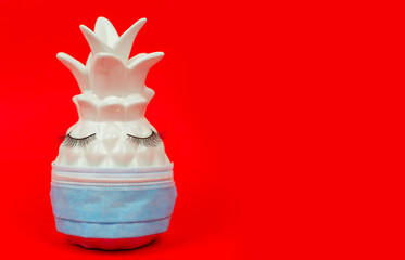 white ceramic pineapple with false eyelashes in protective face mask on red background, minimal creative concept