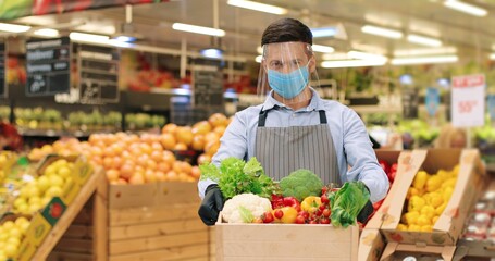 Portrait of handsome Caucasian man in face protection shield standing in food store with vegetables indoors. Male seller in mask holding box with different fresh food in supermarket Quarantine concept