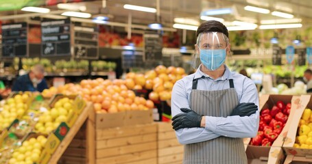 Portrait of handsome young man in face protection shield and apron standing in food store with vegetables indoor. Male Caucasian employee in medical mask looking at camera in supermarket in quarantine