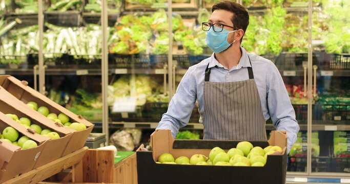 Portrait of Caucasian handsome man worker in mask and apron walking in supermarket with shopping cart with fruits indoor. Male young food store assistant in glasses working in retail. Job concept