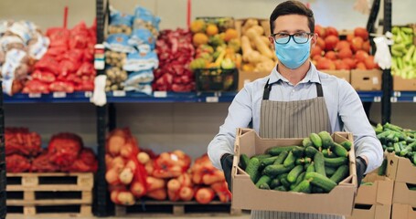 Camera approaching handsome man in face mask standing in food store with vegetables. Male Caucasian employee in glasses holding box with fresh cucumbers in supermarket in quarantine. Close up concept