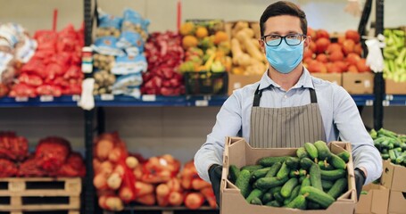 Camera approaching handsome man in face mask standing in food store with vegetables. Male Caucasian employee in glasses holding box with fresh cucumbers in supermarket in quarantine. Close up concept