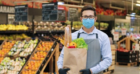 Camera approaching male seller in mask and apron standing in supermarket with packet with food. Close up of handsome Caucasian man worker with vegetables in grocery store alone. Retail concept