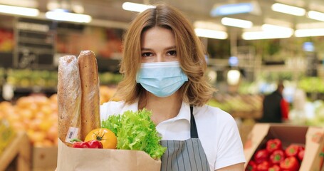 Camera approaching female employee in mask and apron standing in supermarket with vegetables. Close up of beautiful Caucasian woman seller with food in packet in grocery store. Retail concept