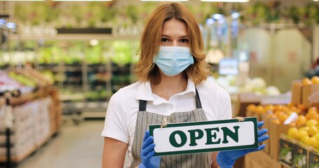 Close up portrait of Caucasian beautiful female seller in medical mask and apron standing in supermarket and holding sign Open. Woman worker in grocery store indoor in quarantine. Employee concept