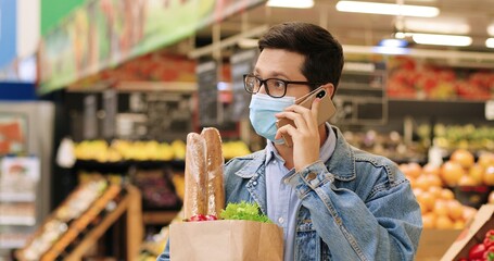 Close up portrait of handsome Caucasian male standing in grocery store with packet with food and talking on smartphone. Man in mask and glasses with vegetables in supermarket calling on cellphone