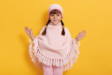 Cheerful little kid girl with pigtails wearing stylish poncho, cap and trousers, poses isolated on yellow background, child spreading hands, looks at camera with happy and excitement facial expression