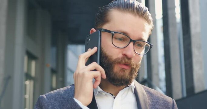 Close up of attractive confident positive 35-aged bearded businessman in glasses which talking on mobile with business partner outdoors on the background of office building