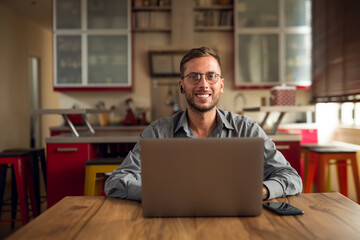 Authentic shot of young successful self confident businessman is smiling satisfied with his work in camera while doing smart working with laptop from home in the kitchen during lockdown. 