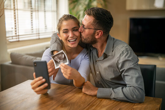 Authentic shot of happy smiling married couple is making video technology call to friends or family with smartphone from home and showing an ultrasound to announce their future child birth.