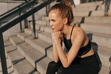European young woman in sport black uniform sitting on concrete staircase. Young woman with fit body running against grey background. Female model in sportswear exercising outdoors.