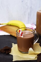 A glass full of delicious cocoa is on the table, with cream on top and chocolate next to it - delicious, sweet chocolate milk on a table, with banana, milk and chocolate