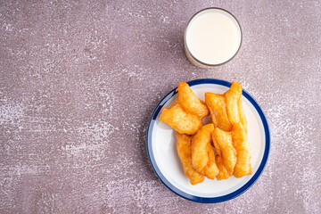 Top view of deep-fried dough sticks or Chinese doughnut sticks on white plate and a glass of soybean milk on gray stone background. Space for text
