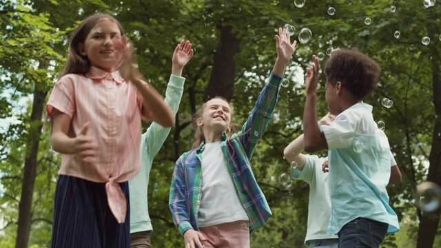 Medium of five multi-ethnic school boys and girls running, jumping in public park, catching and bursting soap bubbles