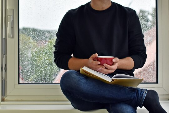 A man in his mid-twenties sits on a windowsill, in the background raindrops can be seen on the window pane - the man is reading a book while drinking a coffee - anonymous picture without head and face - Powered by Adobe
