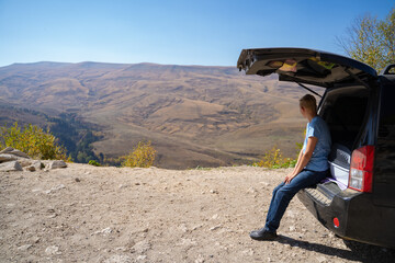 Young man is resting in nature with a car. Uses the trunk of an off-road vehicle for recreation
