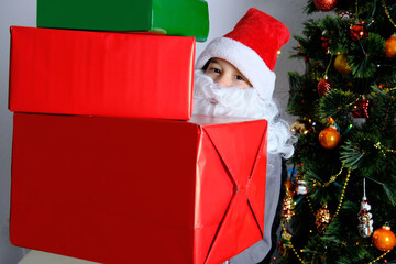 little kid, santa claus helper, an elf with a white beard holds gifts near the christmas tree, the...