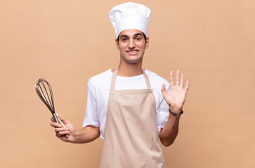 young baker man smiling happily and cheerfully, waving hand, welcoming and greeting you, or saying goodbye