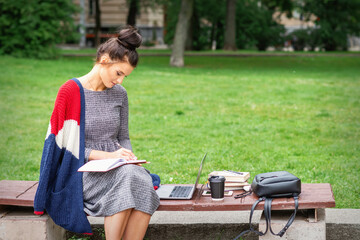 Fototapeta premium Student young woman is writing notes to diary on the bench in the park