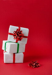 Gifts packed in white paper stand on top of each other, tied with red and green ribbons on a bright red vertical background with copy space