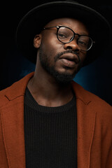 portrait photo of a black man with glasses, black hat, brown jacket and jacket on a dark background in the studio, who is looking thoughtfully at the camera