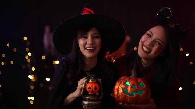 An asian woman wearing a witch costume holds an orange pumpkin. Young women were at the Halloween party. Two girl are having fun at the Halloween party.