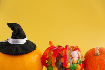 A pumpkin in a witch hat, a jar of jelly worms and colorful candies on a yellow background