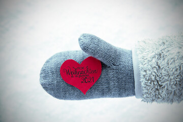 Fototapeta premium Red Heart With German Calligraphy Frohe Weihnachten Und Ein Glueckliches 2021 Means Merry Christmas And A Happy 2021. Human Hand In A Glove With Fleece And Snow.
