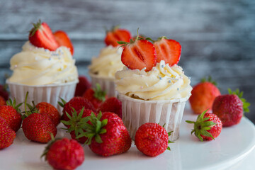 cupcake in the center with white cream decorated with ripe strawberries on a gray blue background and scattered strawberries.
