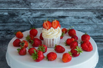 cupcake in the center with white cream decorated with ripe strawberries on a gray blue background and scattered strawberries.
